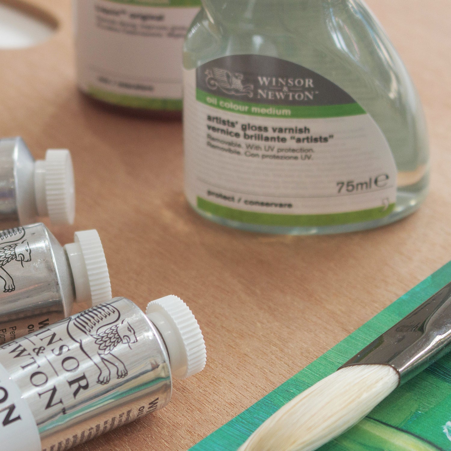 Close up of a desk featuring Winsor & Newton's gloss varnish, oil paint tubes and a hog hair paintbrush.