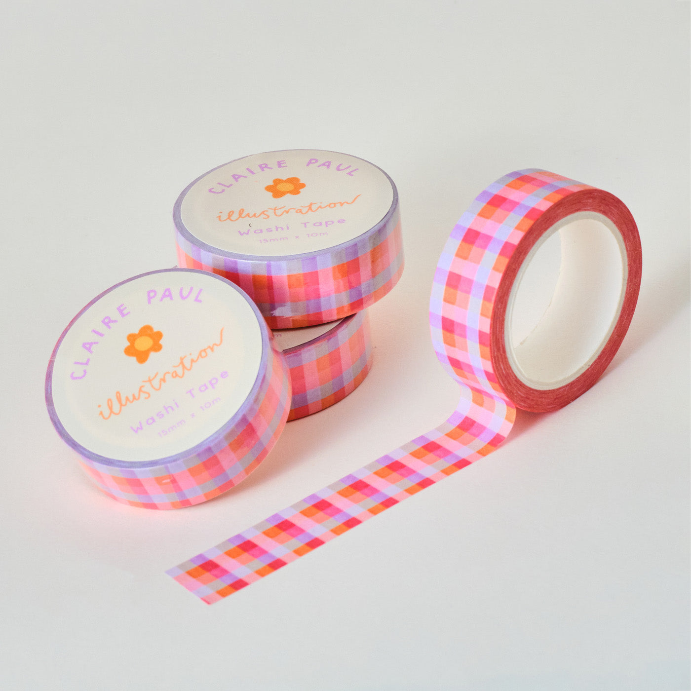 Stack of Pink Gingham washi tapes in their packaging with an open roll. Tapes feature a gingham pattern in pink and lilac. Each roll measures 15mm wide and has 10m of tape. Made by Claire Paul. 