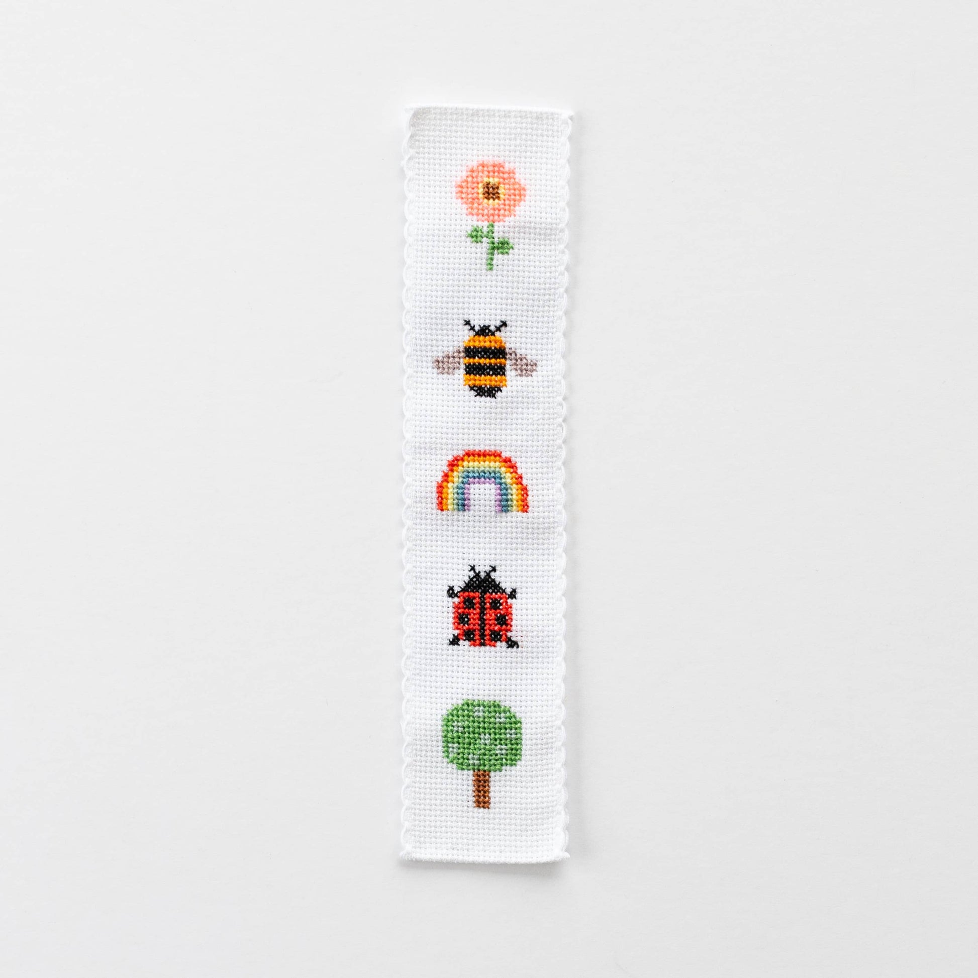 A finished cross-stitched bookmark with five nature motifs including a flower, bumblebee, rainbow, ladybird and tree.