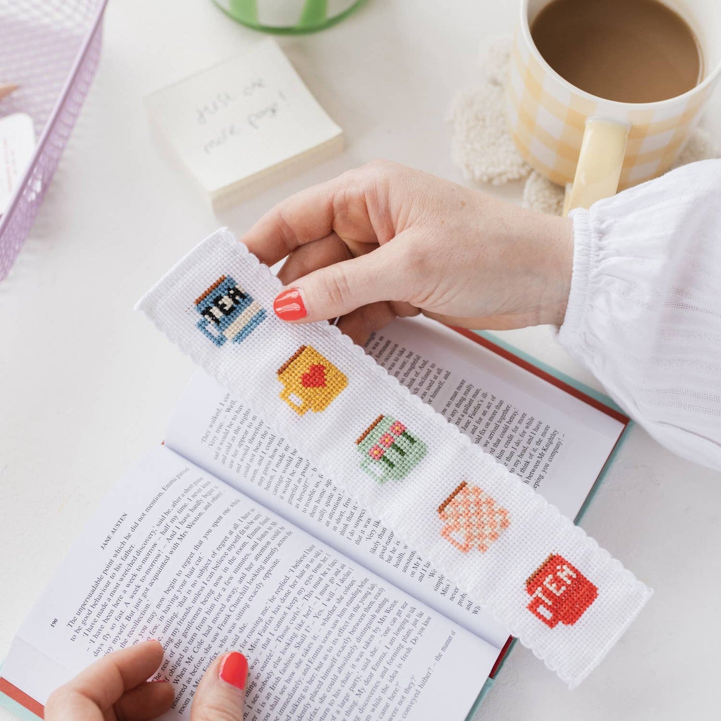 A finished cross-stitched tea mug patterned bookmark marking a place in a book.