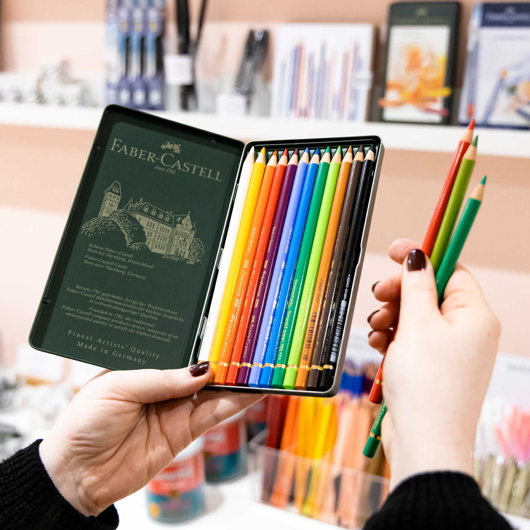Person holding the green metal tin of 12 polychromos coloured pencils in front of a shelf of art materials in Salt Art Supply. 