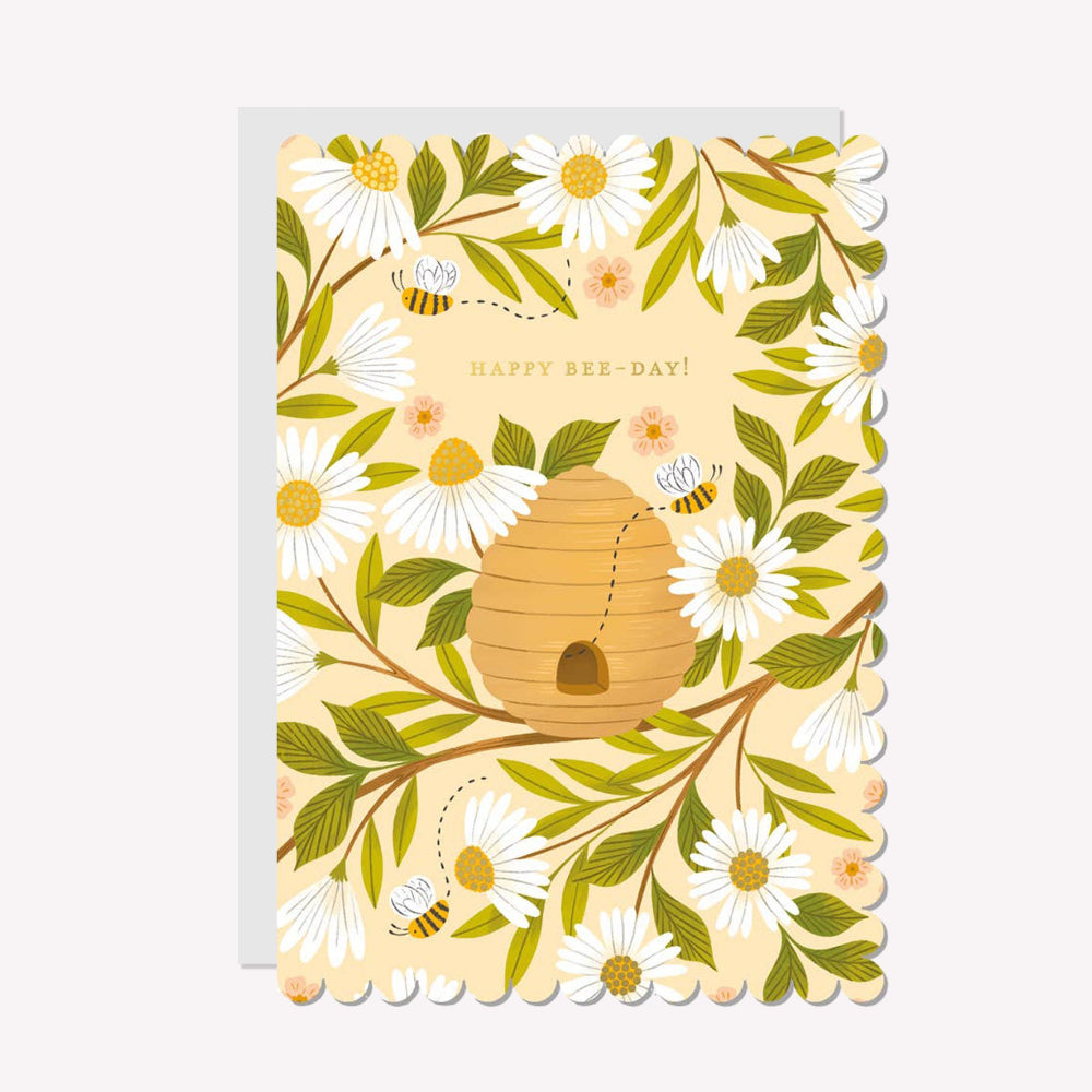 An illustrated birthday card by Raven Paper Co, printed by Ricicle Cards. This card features a hive with several bees buzzing around, with gold-foiled text above that reads “Happy Bee-Day!".