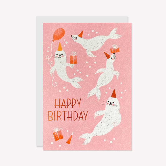 An illustrated birthday card by Ricicle Cards. This card features four swimming seals with party hats printed onto a pink background with text that reads "Happy Birthday".