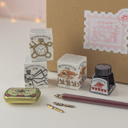 close up photo of contents of the Salt Art Supply, Calligraphy for Beginners gift set including three drawing inks, a dip pen, calligraphy nibs and a nib storage tin, packaged in a festive branded box for Christmas gifting. 