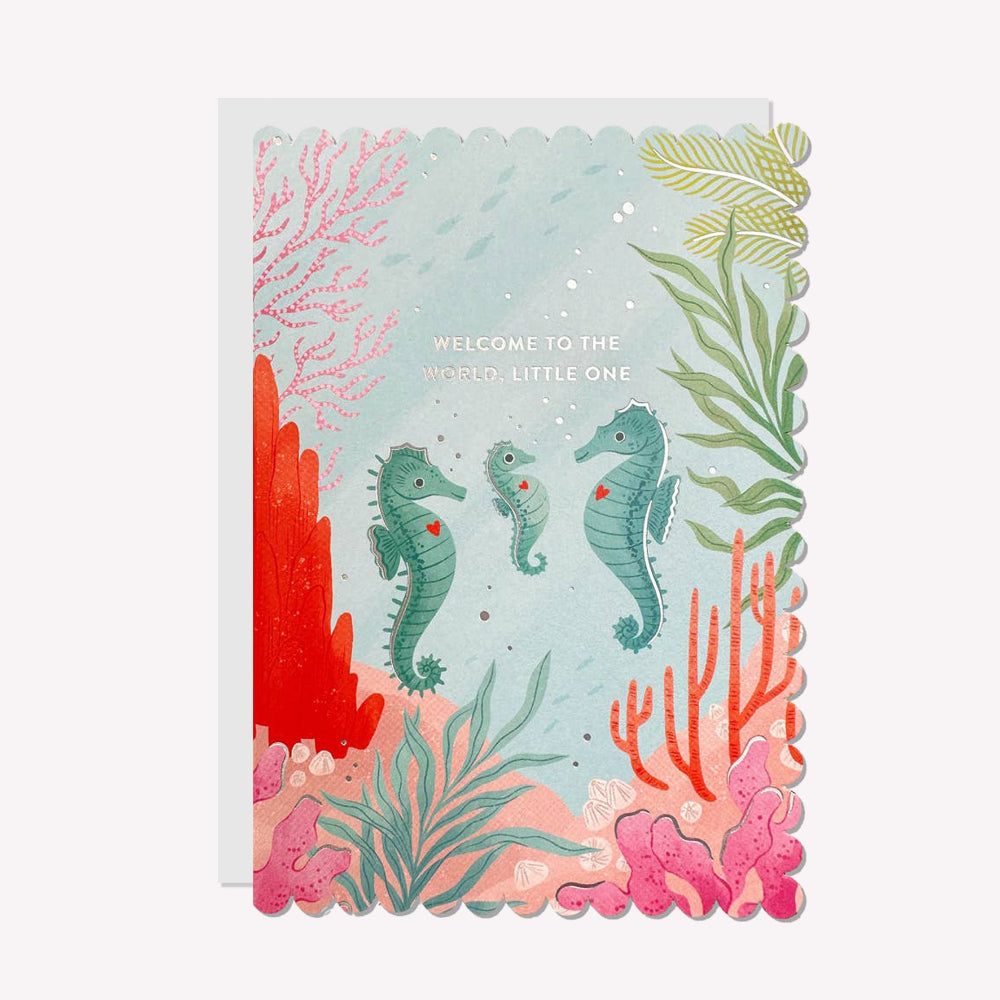 An illustrated new baby card by Raven Paper Co, printed by Ricicle Cards. This card features a family of seahorses surrounded by coral, with silver-foiled text above that reads “Welcome To The World, Little One".